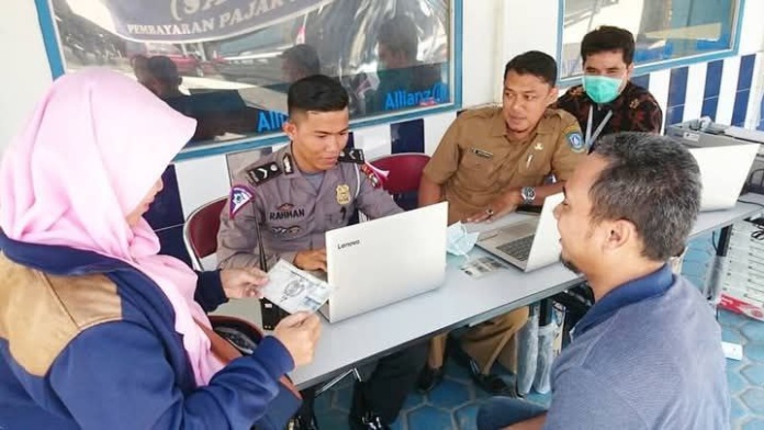 Sekelompok orang sedang melakukan proses administrasi di sebuah meja layanan publik. Seorang petugas berseragam polisi duduk di depan laptop, berhadapan dengan seorang wanita berhijab yang menyerahkan dokumen. Di sebelahnya, seorang petugas berseragam cokelat turut membantu di depan laptop lain, sementara seorang pria berpakaian biru duduk di sisi meja yang sama. Di belakang mereka, terlihat satu orang lagi mengenakan masker hijau dan pakaian batik, tampak sedang mengamati proses tersebut. Latar belakang menunjukkan papan dengan logo 